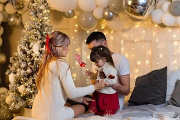 Time To Celebrate Christmas And Give Gifts. Young Family Against Background Of Christmas Lights And New Year Tree. Little Girl Holding Lollipop. Celebration Atmosphere