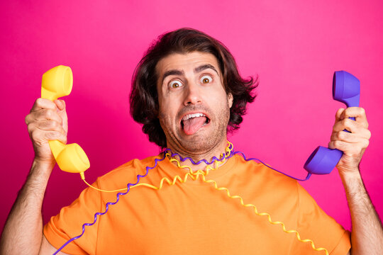Photo of funky guy tongue-out arms hold two telephones hang cables wear orange t-shirt isolated pink color background