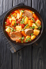 Thick stew of potatoes carrots peas beef tongue and tomatoes close-up in a bowl on the table. vertical top view from above