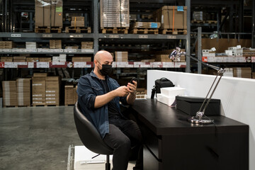 Man in mask sitting in large warehouse office is looking at his mobile phone.