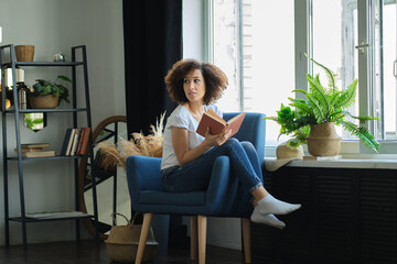 Beautiful young African American woman reading a book lying comfortably on her back on a armchair in the living room in her jeans