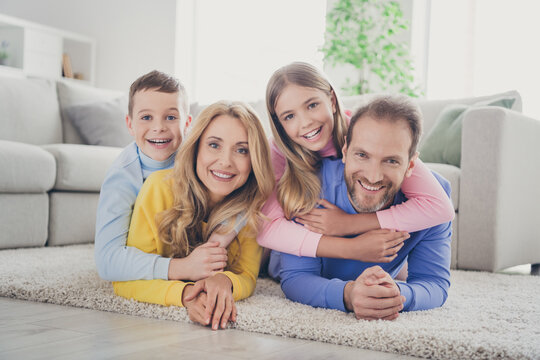 Photo of positive cheerful mommy daddy lying carpet hug two kids boy girl piggyback in house indoors