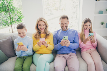 Full body photo of positive family people sit sofa mom dad two small kids use smartphone in house indoors