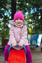 Little blonde girl in a pink coat and knit hat rides on a swing at the playground. Four-year-old girl laughs. Entertainment in the autumn park in the cold season. Сoncept of a happy childhood