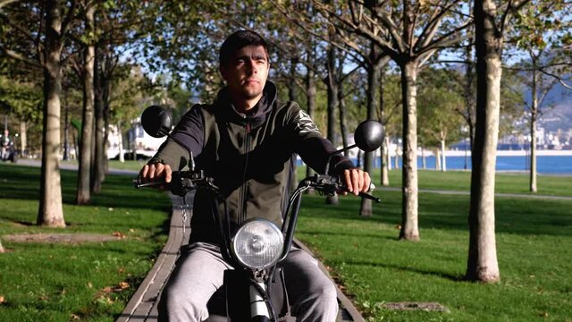 Young man on electric scooter is riding through publick park at sunny morning, front view steadicam shot. Ecological electro transport concept.