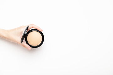 A female hand holds a compact beige powder on a white background, top view. Makeup nude product for...