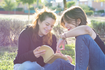 Two smiling young women reading a book sitting on the green grass