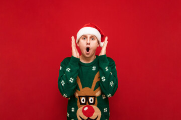 Portrait of shocked young man in christmas clothes on red background, looks into camera with mouth open and screams. Amazed guy in santa hat isolated on red background.