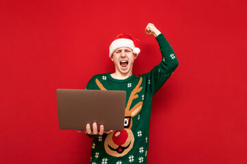 Joyful young man in Christmas clothes stands with a laptop in his hand and is emotionally pleased to win on a red background, looks into the camera with his arm raised up and shouts with happiness.