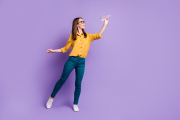 Full length profile photo of girl hold paper plane empty space wear specs yellow shirt blue trousers sneakers isolated violet color background