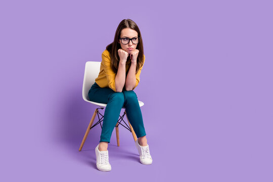 Full length photo of lady sit chair arms chin look empty space wear yellow shirt blue pants specs sneakers isolated purple color background