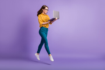 Full length profile photo of lady jump hold netbook look screen wear yellow shirt blue pants sneakers isolated violet color background