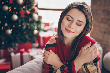 Photo of charming lady closed eyes wrapped checkered plaid hug herself wear red sweater in decorated x-mas living room indoors