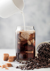 Pouring milk into glass with iced black coffee with glass jar of coffee beans and salted caramel on light background.