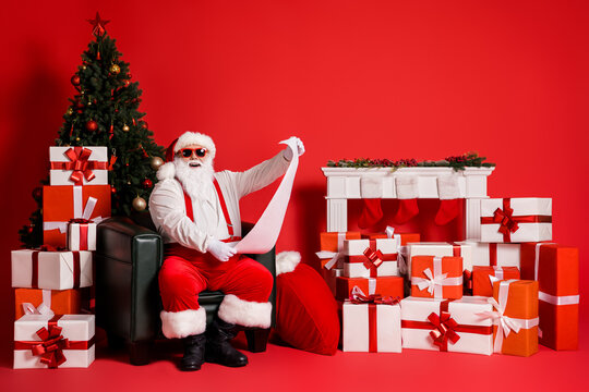 Portrait of his he attractive funky fat cheerful cheery Santa sitting in armchair reading long paper letter giftbox pile stack order North Pole isolated bright vivid shine vibrant red color background