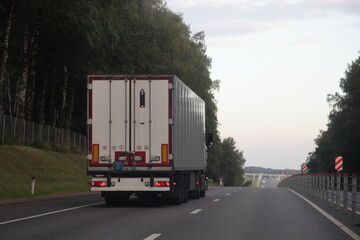 European heavy truck with white semi trailer van drive on left side of two lane suburban asphalted one way road, rear view at summer evening on forest and sky background, transportation logistics © Ilya