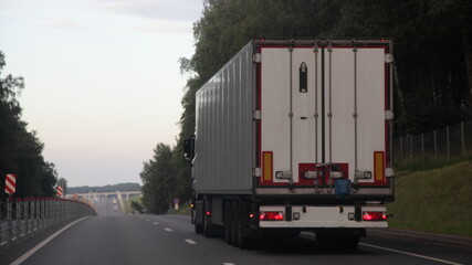 European heavy truck with white semi trailer van drive on two lane suburban asphalted highway road, back view at summer evening on forest and sky background, transportation logistics