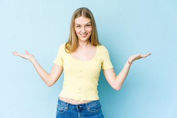 Young blonde woman isolated on blue background makes scale with arms, feels happy and confident.