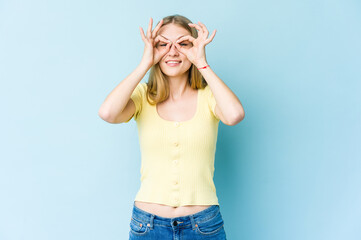 Young blonde woman isolated on blue background showing okay sign over eyes