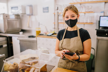 young blonde-haired barista owner wearing a mask and checking stock in a coffee shop with a smartphone in her hand, and looking to he camera