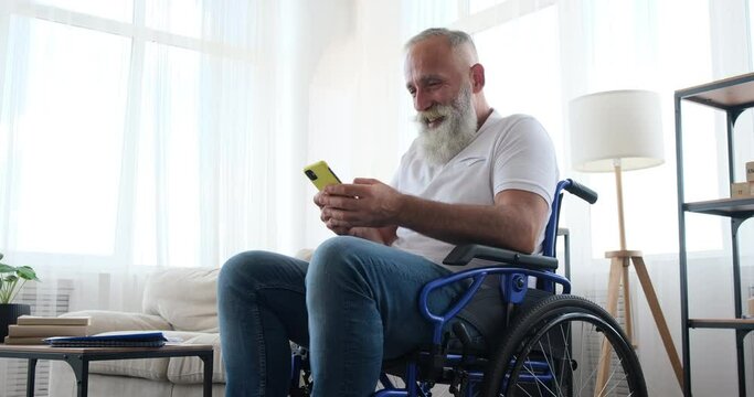 Disabled old man having fun text messaging using mobile phone sitting on wheelchair at home