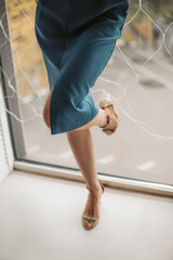 woman in blue dress and high heels stands at the window with her heel against the glass