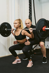 Fototapeta na wymiar Young muscular woman doing barbell squats with personal trainer. Private crossfit workout in a small fitness studio