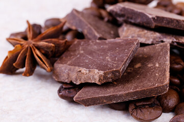 Stack of chocolate bar pieces with heap of roasted coffee beans and spices, cinnamon sticks and anise stars on a white table. Macro shot.