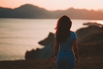 Picturesque mountain landscape at sunset with the sea on the horizon, a young beautiful woman walks down the slope in the orange rays of the sunset