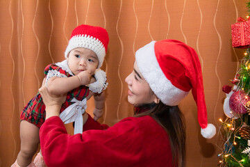 Asian happy young mother and adorable baby on sweater clothes sit near Christmas tree decoration play together at home in Holiday greeting season, beautiful mom with Santa red hat kissing sweet girl