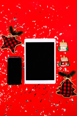 Tablet and phone with Christmas or New Year ornaments on red background with confetti. Red festive backdrop. Buffalo plaid ornaments. Holidays at home concept. Virtual greetings. Stay home