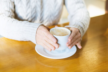 Close-up of a woman's hands in a warm sweater holding a white Cup of hot coffee, chocolate or tea in a coffee shop against the background of a wooden table. The concept of winter comfort, holiday.