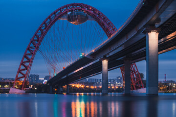 View of Zhivopisny Bridge in Moscow, Russian Federation. Photo shoot of the red arch, steel cable-stayed bridge over the Moskva River. Evening. City lights and bridge lights are reflected in the water