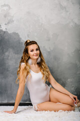 Beautiful young girl in swimsuit with feathers. Stylish woman in boho style bodysuit sits on floor on gray wall background.