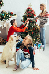 selective focus of kid in sweater cuddling labrador near parents decorating christmas tree in living room