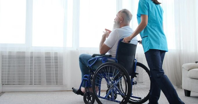 Female nurse pushing old man on wheelchair and having a discussion looking out through window