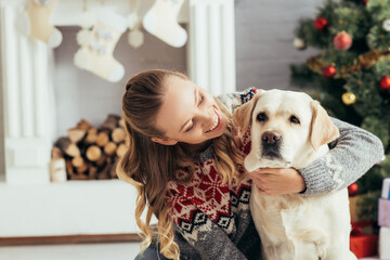 happy woman in sweater looking at labrador near christmas tree and fireplace