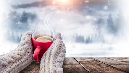 Red cup with coffee, tea in female hands on a snowy background, winter forest. A red cup in hands against the background of a winter forest landscape on soft, knitted sleeves. Coffee, tea on a wooden 