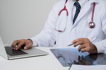 Close up of Asian doctor put on stethoscope working on laptop with x-ray film sitting at the desk isolated on white background.