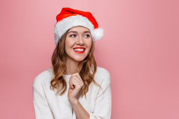 Christmas girl. Young caucasian woman in santa hat and white sweater smiling and looking at copy space isolated on pink background