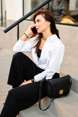 Handsome smiling woman with mobile phone sitting near restaurant. She looks suprised. She wears white shirt and black trousers, because she is young business woman.