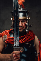 Warlike armoured roman warrior with red mantle posing holding his sword with serious face in dark...