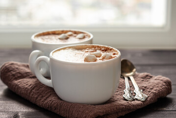 Two white cups of cappuccino with marshmallows and spoons on wooden table. Close up photo with selective focus