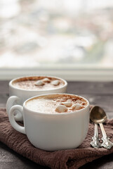 Two white cups of cappuccino and two spoons, lying near the cups on a brown cloth. Close up photo with selective focus