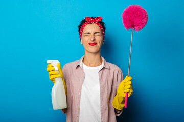 Screwing up is eyes young woman in gloves holding spray and dust brush on blue background.