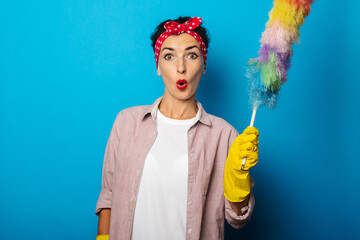 Surprised young woman in gloves holding dust brush on blue background.