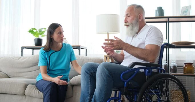 Female home caregiver talking to disabled old man sitting on wheelchair at home