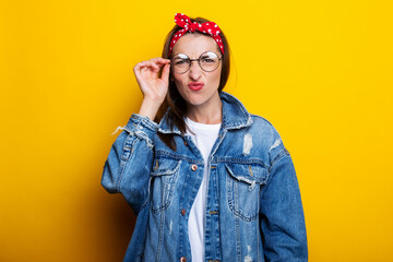 Young woman in a hair band, in a denim jacket with glasses closed her eyes on a yellow background.