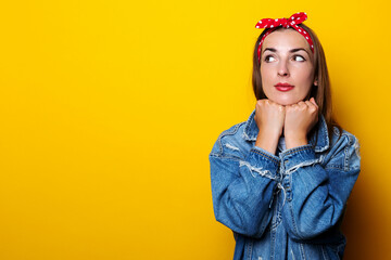 Obraz na płótnie Canvas Young woman in a hair band, in a denim jacket, holds her hands under the edge and looks to the side on a yellow background.