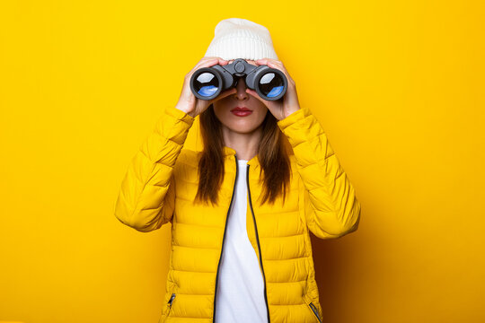 Young woman in a yellow jacket in a hat looks through binoculars on a yellow background.